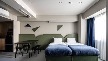 A modern hotel room with a bed, a desk, and chairs. There is a kitchenette in the corner.