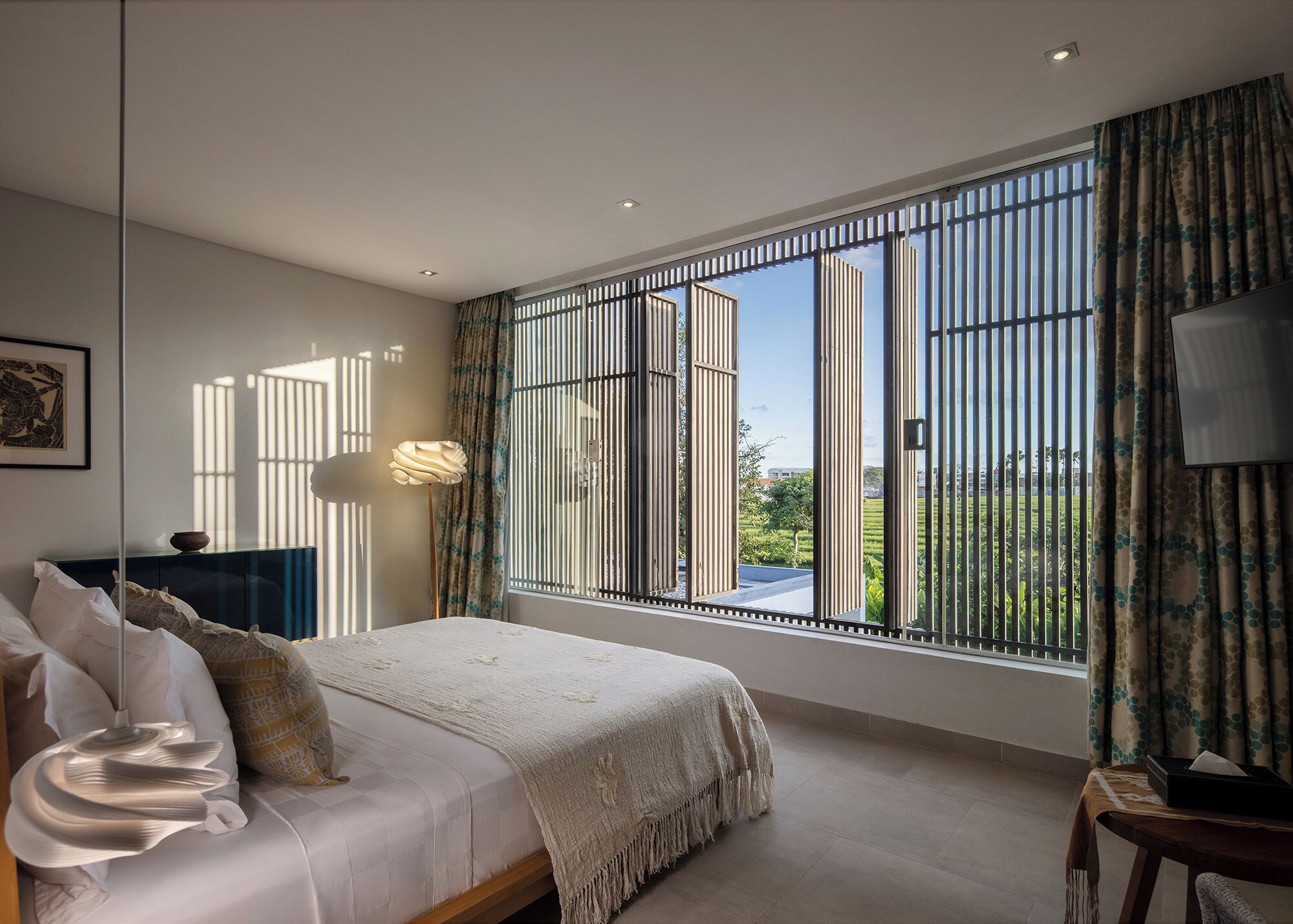 Quarto com uma janela grande, uma cama, mesa de cabeceira, abajur e televisão.