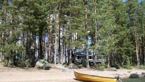 Uma cabana rodeada por altas árvores de pinheiro numa praia arenosa, com um barco amarelo na água.