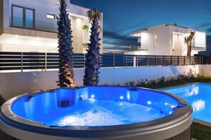 A swimming pool with a hot tub, surrounded by a modern house with a balcony and palm trees.