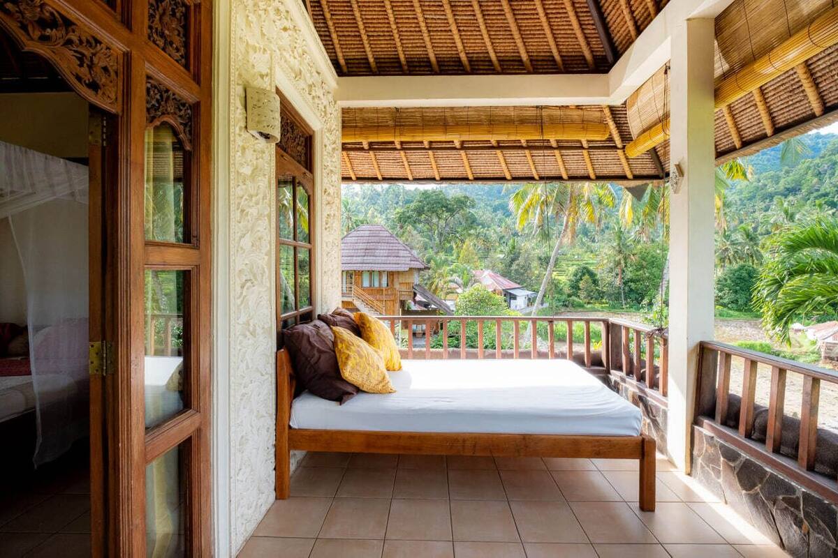 Una cama con estructura de madera y colchón blanco, un balcón con barandilla de piedra y vistas a un paisaje tropical con palmeras y una casa al fondo.