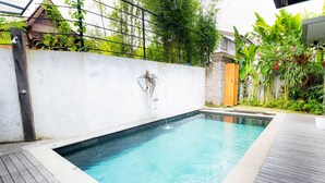 Una piscina rectangular amb un solàrium de fusta al voltant, una paret blanca amb una escultura metàl·lica, i vegetació abundant al fons.
