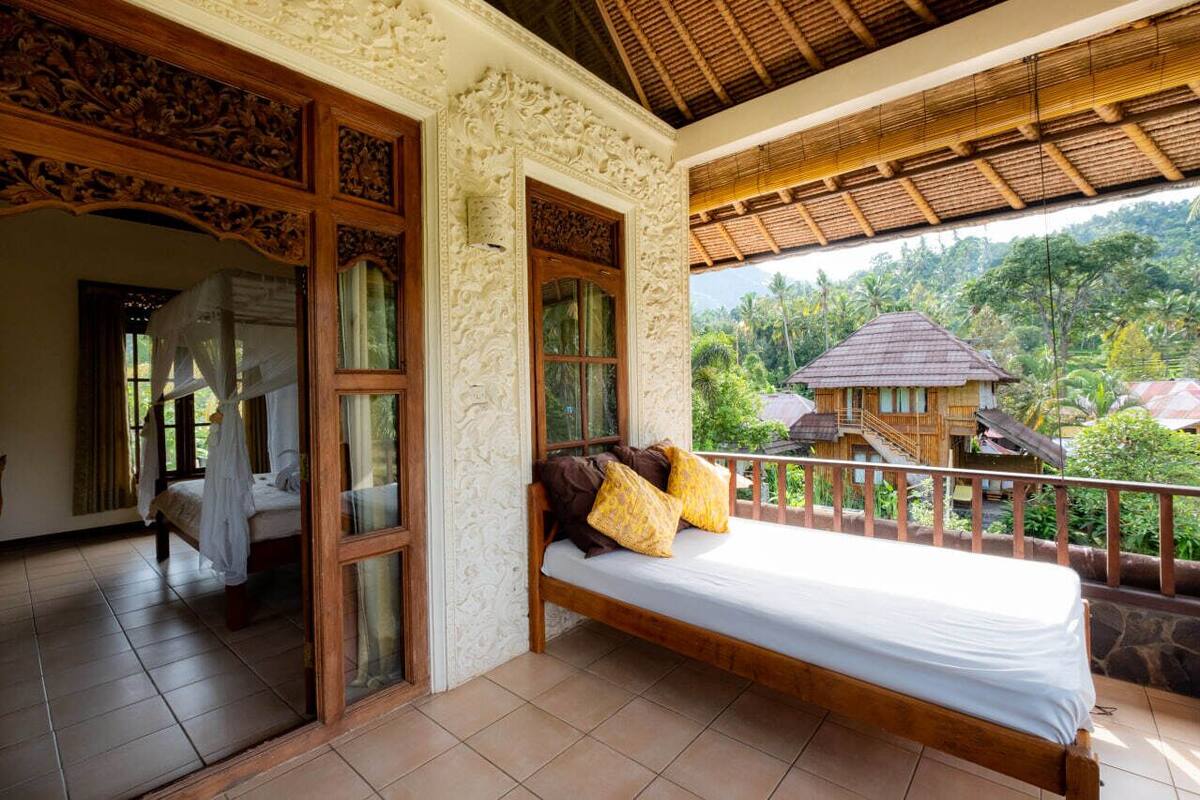 Una habitación con una cama, un balcón con vista a una casa y árboles, y techo de madera.