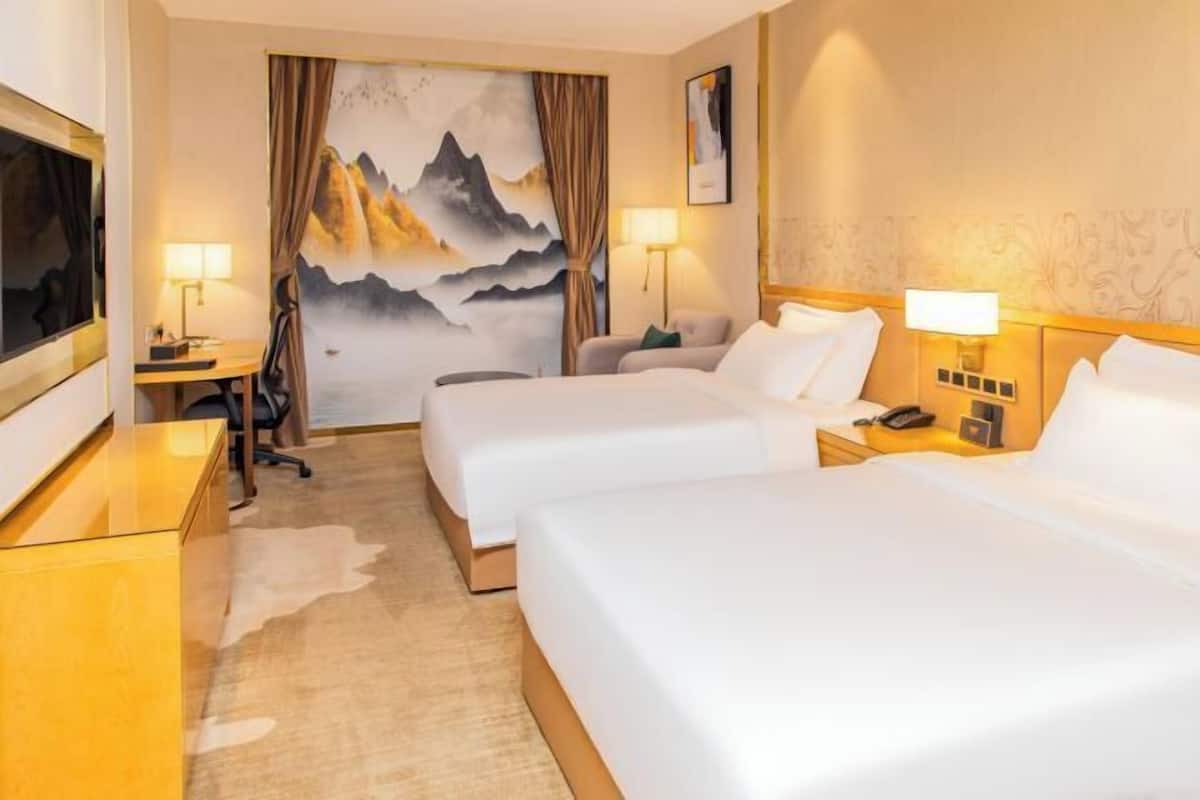 A hotel room with two beds, a desk, a chair, and a large painting of mountains.