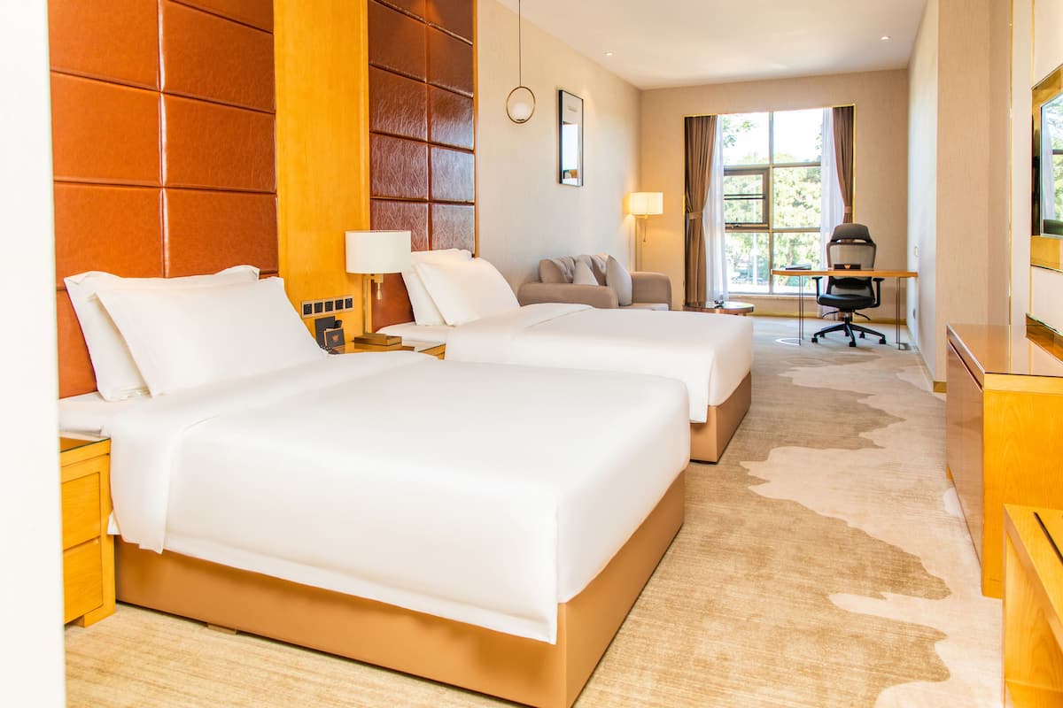 A hotel room with two beds, a desk, a chair, a lamp, and a large window.