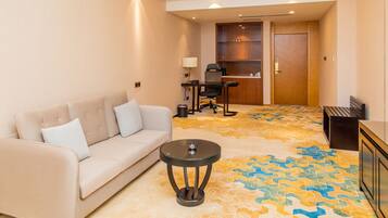 Habitación de hotel con sofá color crema, una mesita redonda pequeña, un escritorio con una silla y una puerta de madera.