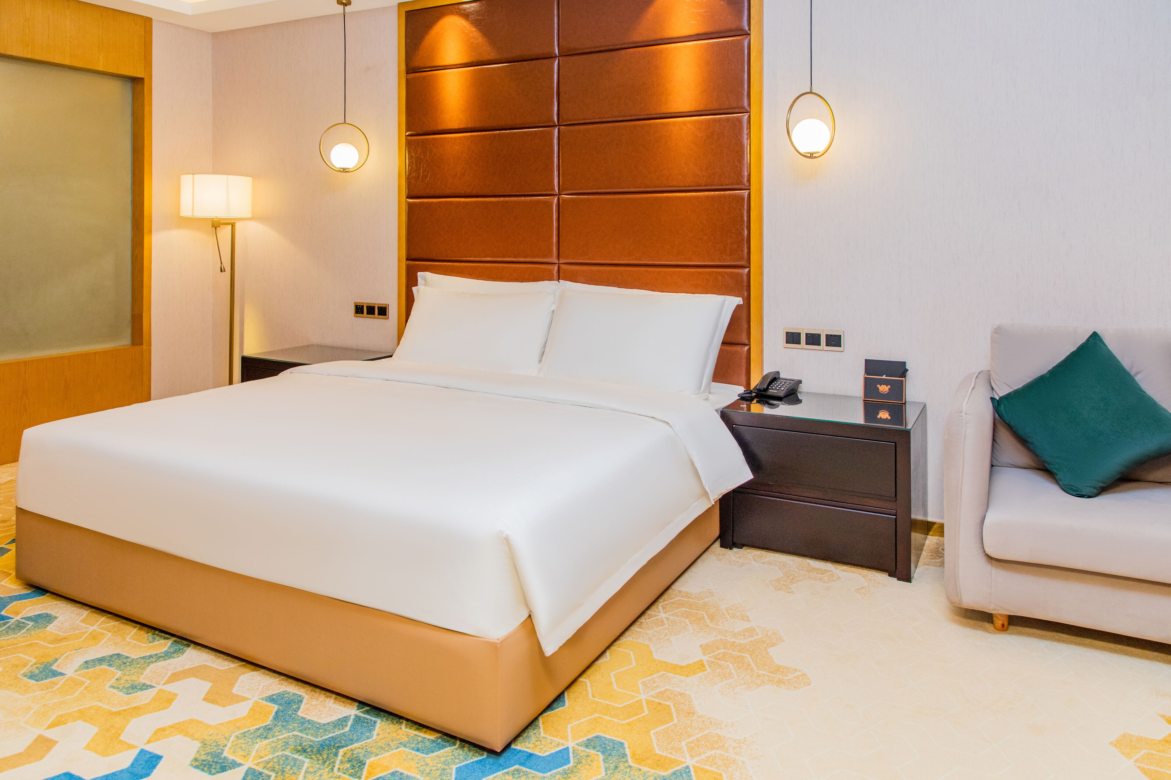 A hotel room with a large bed, a nightstand, a lamp, a sofa, and a patterned carpet.