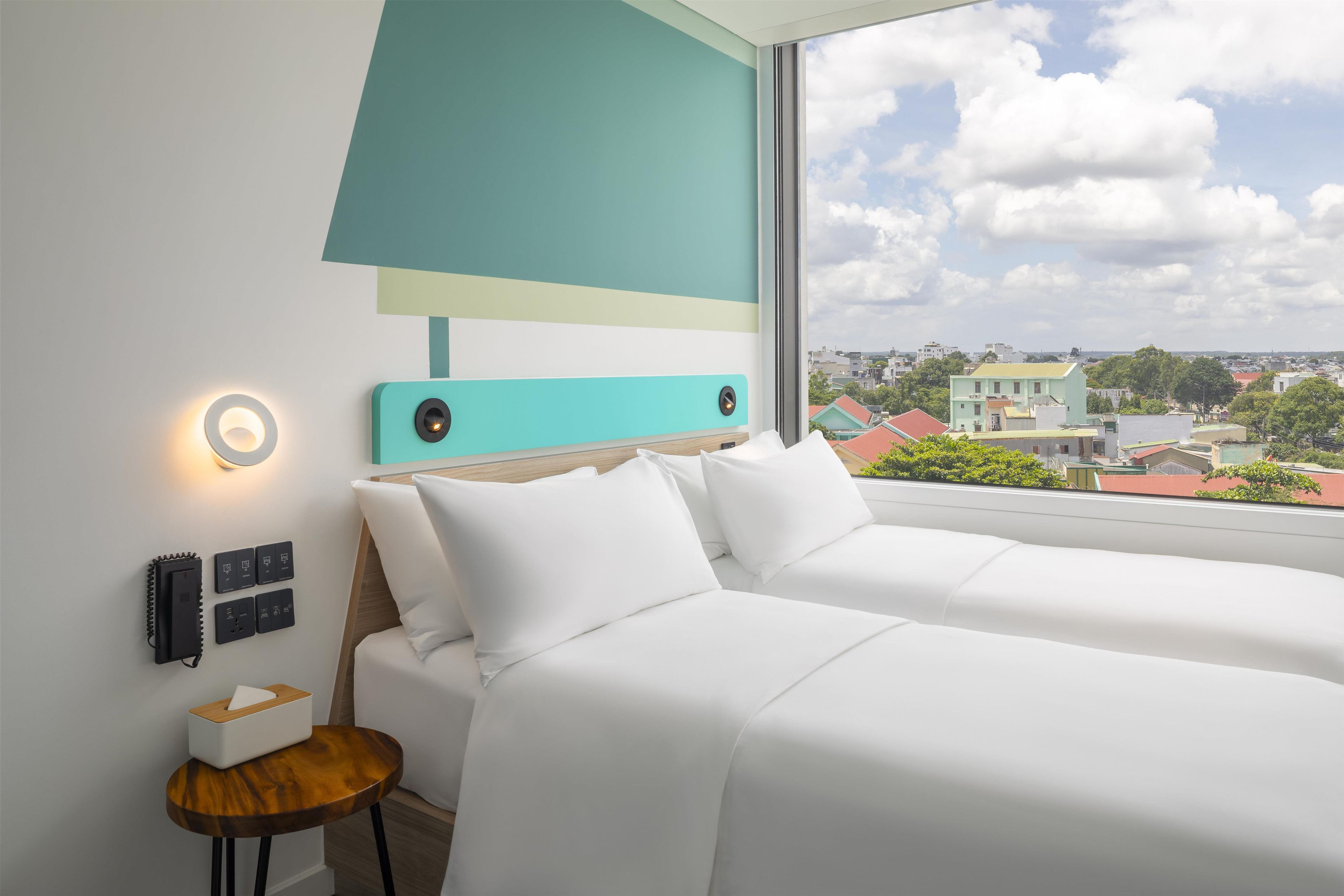 Ein Hotelzimmer mit großem Fenster, einem Bett, einem Nachttisch und einem an der Wand befestigten Telefon.