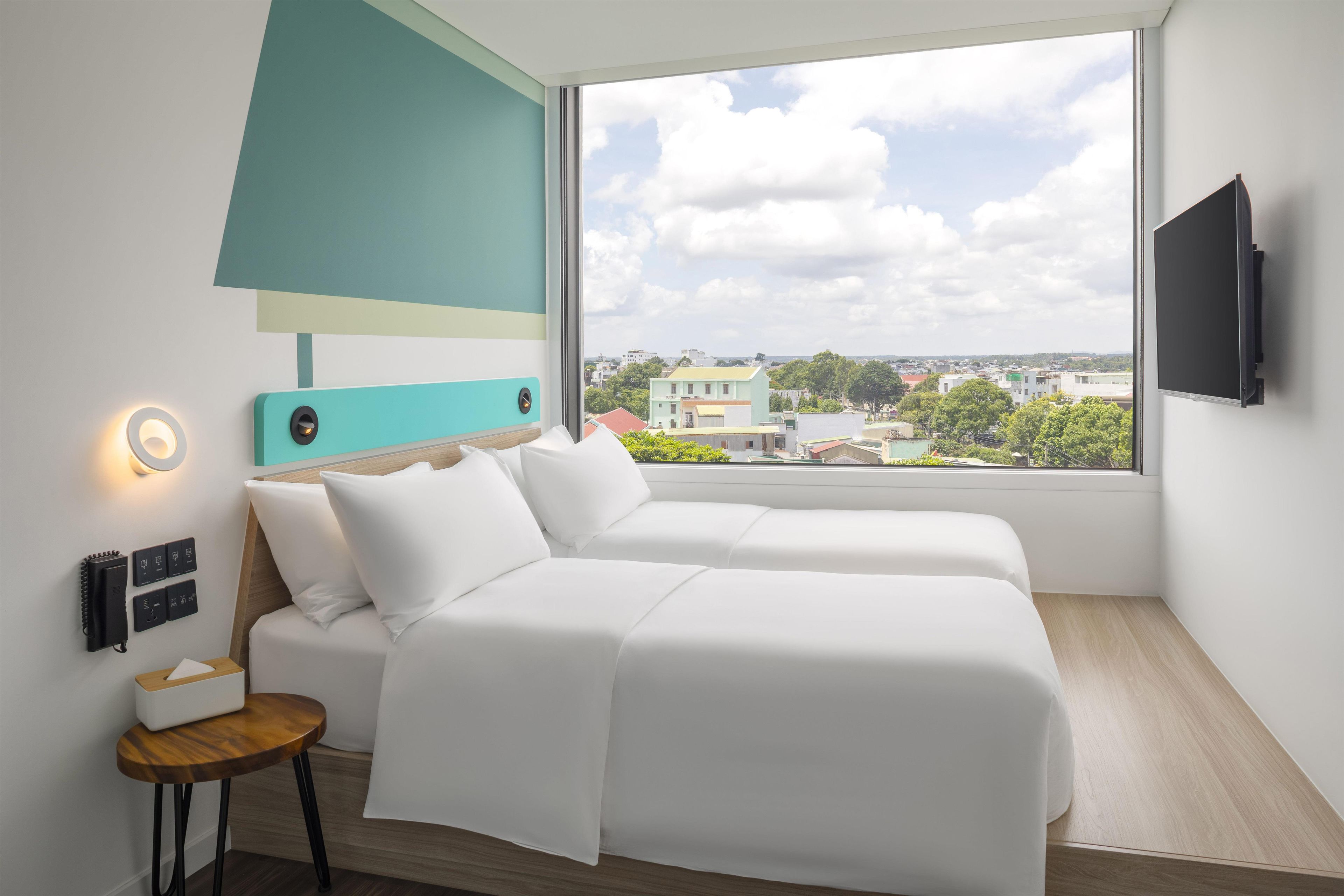 Una habitación de hotel moderna con un ventanal grande, una cama, una mesita de noche y un televisor montado.