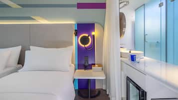 A modern hotel room with a bed, a bedside table, a mirror, a small table with a tissue box, a minibar, and a bathroom with a sink and mirror.