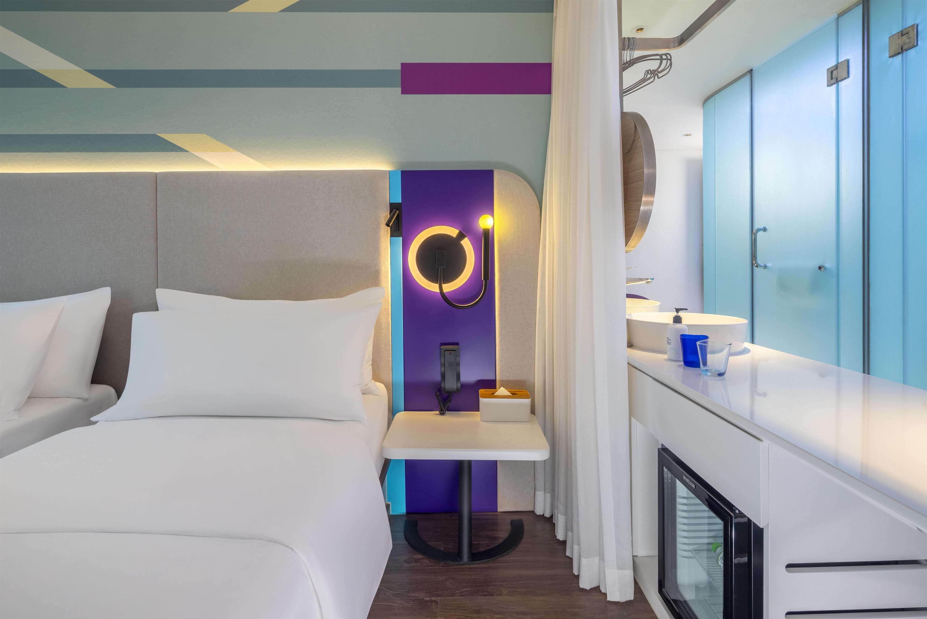 A modern hotel room with a bed, a bedside table, a mirror, a small table with a tissue box, a minibar, and a bathroom with a sink and mirror.