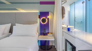 A modern hotel room with a bed, a bedside table, a mirror, a small table with a tissue box, a minibar, and a bathroom with a sink and mirror.