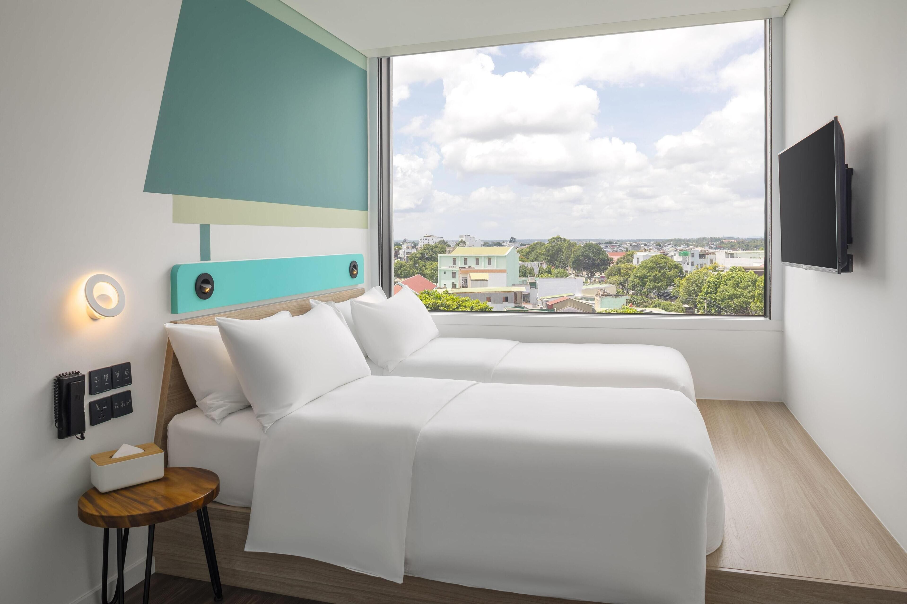 A modern hotel room with a large window, a bed, a bedside table with a tissue box, a wall-mounted TV, and a small lamp.