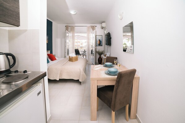 A compact hotel room with a kitchenette, a bed with a red pillow, a dining table with blue plates, and a chair.