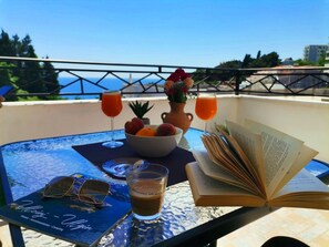A table with a book, two glasses of orange juice, a bowl of fruit, a cup of coffee, and sunglasses on a balcony overlooking the sea.