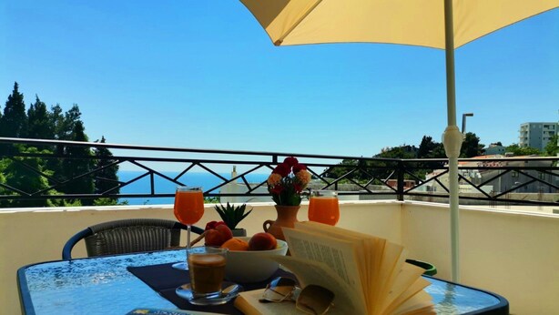 Une table avec un livre, des lunettes et des boissons sur un balcon donnant sur la mer.