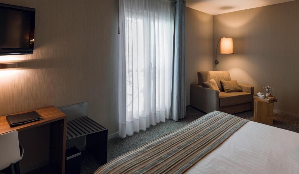 A hotel room with a bed, a desk, a chair, a TV, and a window with curtains.