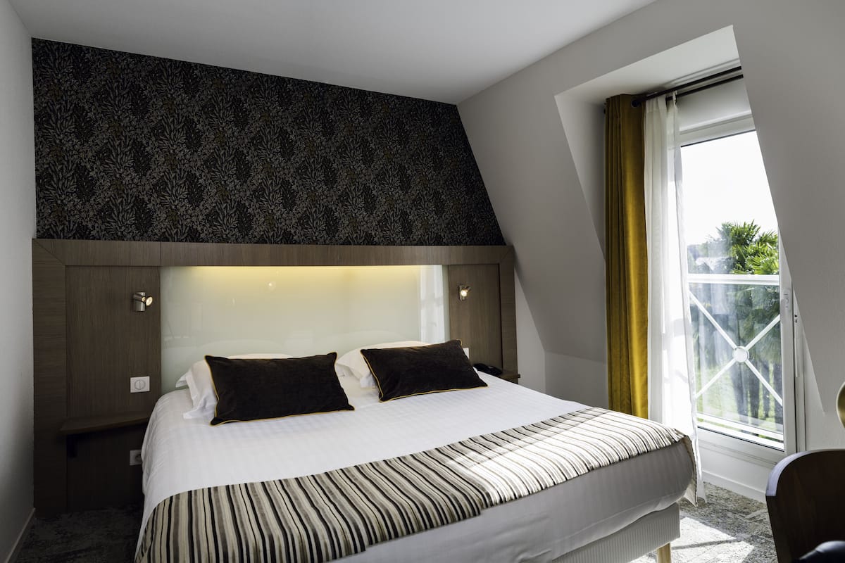 A modern hotel room with a large bed, a dark patterned headboard, and a view of greenery through a window.