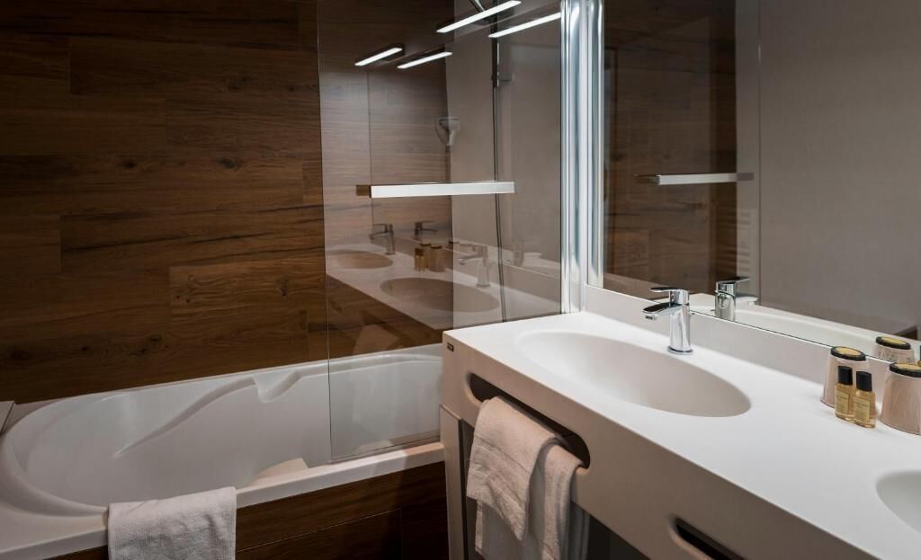 Un baño moderno con ducha envuelta en vidrio, lavamanos blanco con un espejo grande y paneles de madera en la pared.