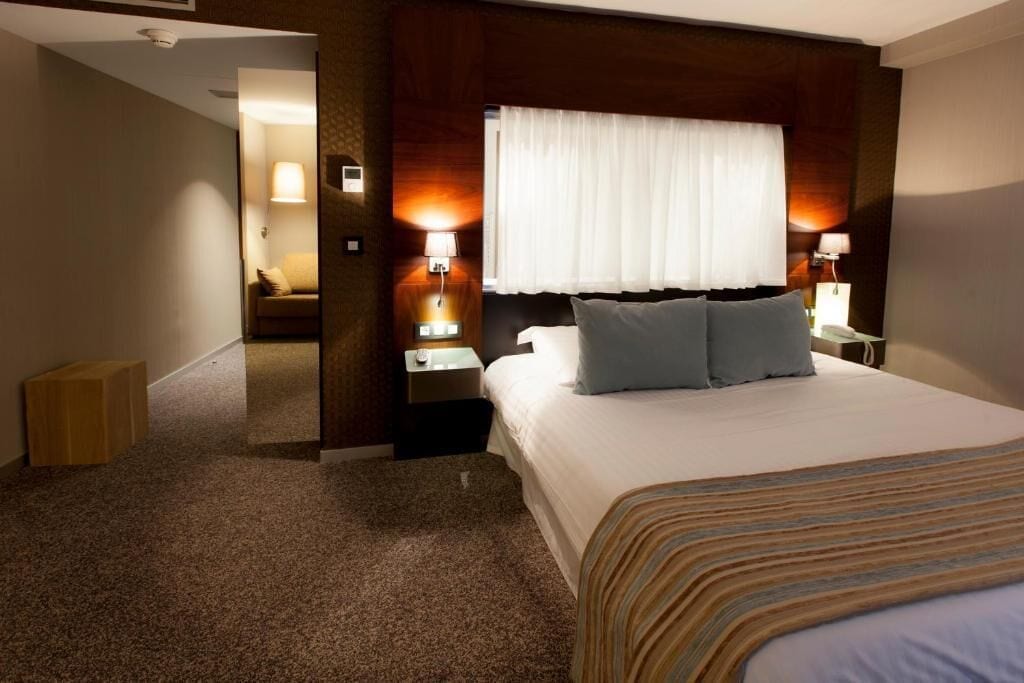 A hotel room with a large bed, two bedside lamps, a window with curtains, and a carpeted floor.