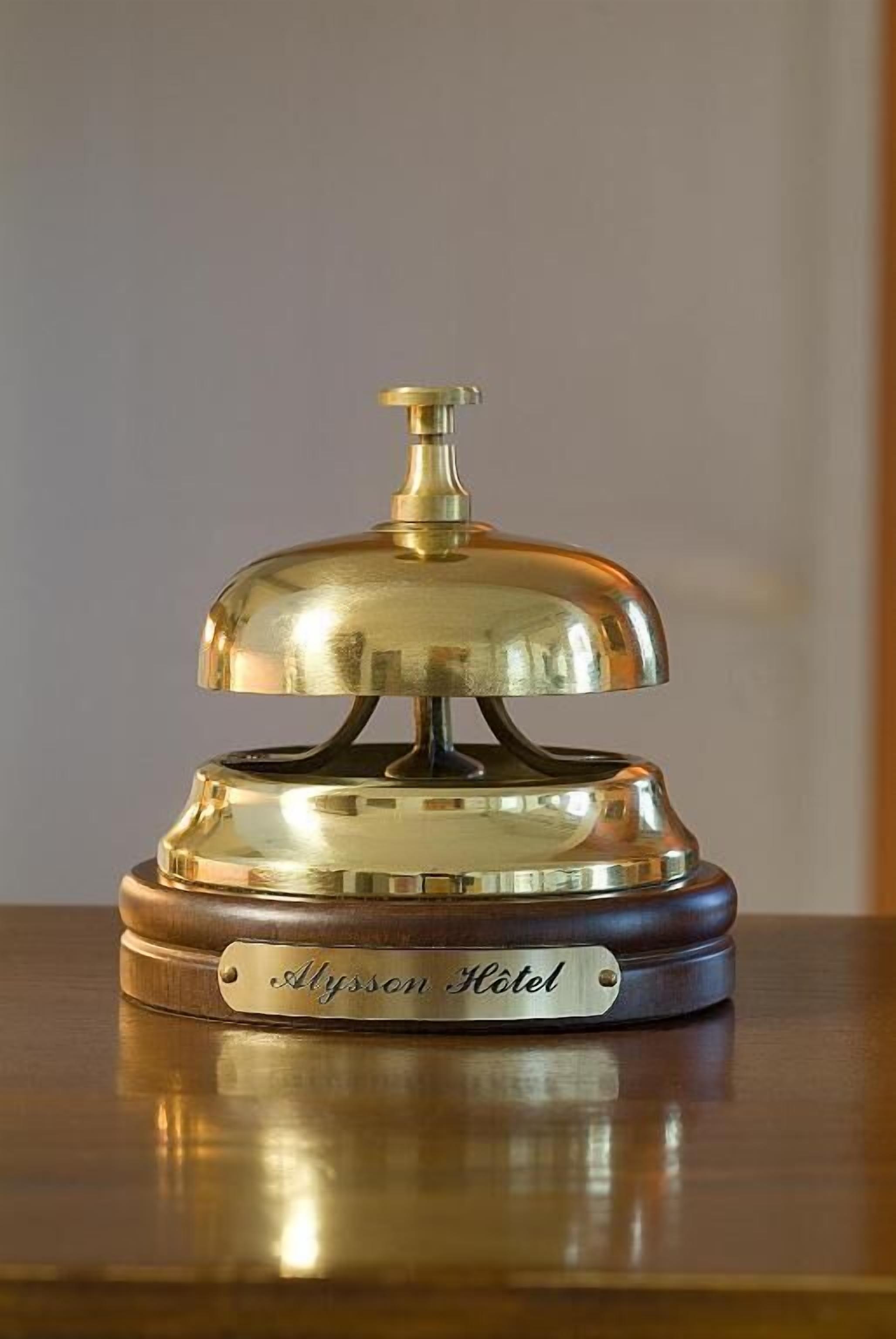 Een bronzen hotelbedieningsbel met een label waarop 'Alysson Hotel' staat, op een houten tafel.