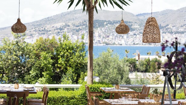 An outdoor dining area with tables and chairs, a palm tree, hanging wicker lamps, and a view of a city and sea. - Harmony Hotel (Sarandë)