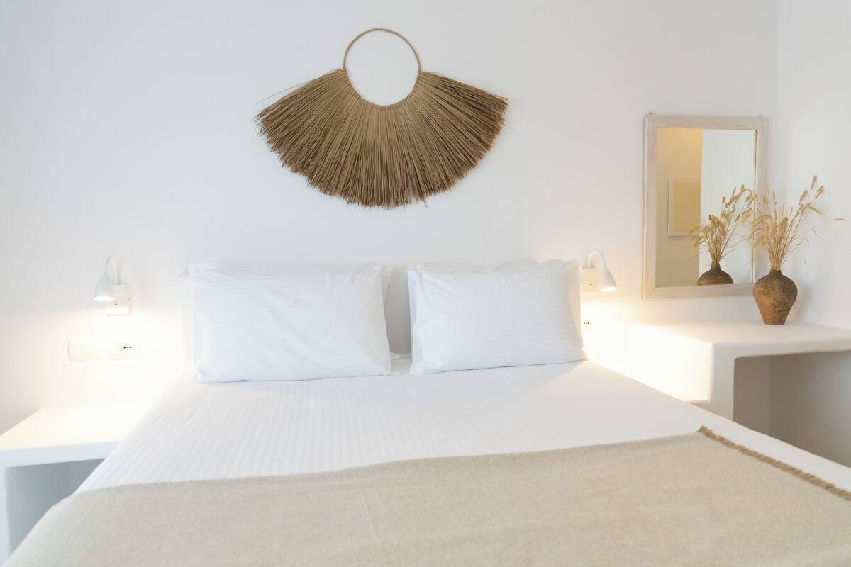 A bed with white bedding, a wall-mounted fan, bedside lamps, and a mirror with a vase of dried plants.