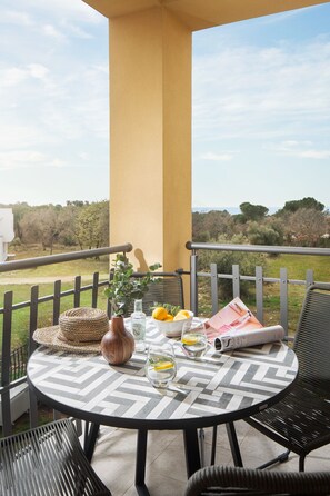 A round table with a patterned tablecloth, a woven basket, a wooden vase, a glass with a lemon, and a rolled-up magazine on a balcony with a view of trees and a fence. - Modern apartment with hot tub (Poreč)