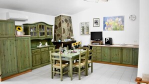 Une cuisine avec des meubles de couleur verte, une table à manger avec des chaises, une cheminée en pierre, un téléviseur et une horloge au mur.