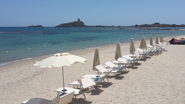 Una playa con filas de tumbonas y sombrillas, un mar azul cristalino y una isla distante.