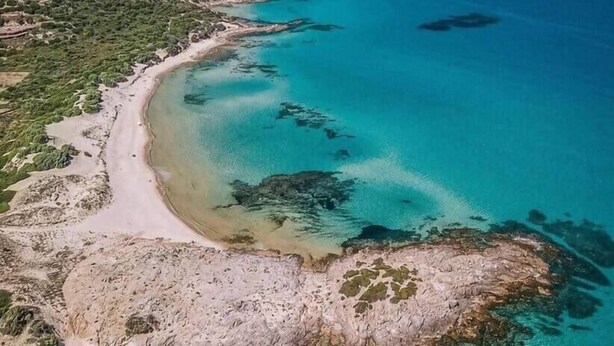An aerial view of a coastal area with a beach, clear waters, and surrounding hills.