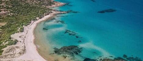 Vue aérienne d’une zone côtière avec une plage, des eaux cristallines et des collines environnantes.