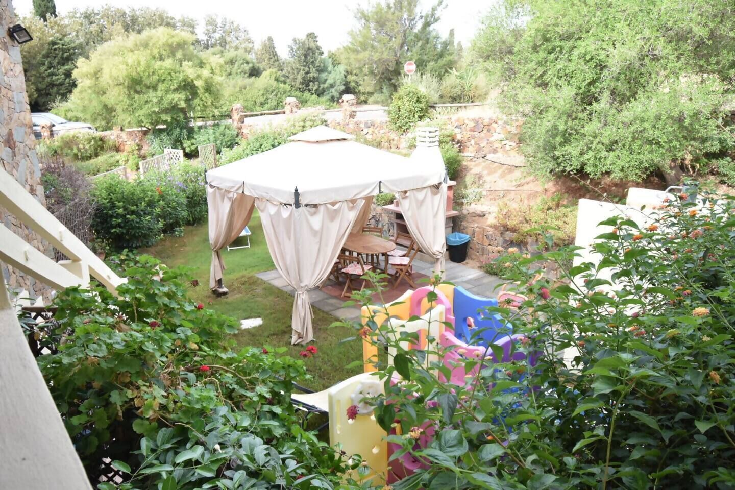 Un área de estar al aire libre con techo, con una mesa y sillas, rodeada de follaje y un muro de piedra.