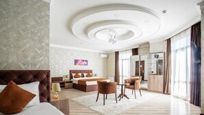A hotel room with a large bed, a sitting area with a table and chairs, a minibar, and a view of the outside through large windows. - Manifa Hotel (Tashkent)