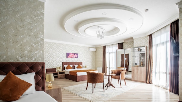A hotel room with a large bed, a sitting area with a table and chairs, a minibar, and a view of the outside through large windows. - Manifa Hotel (Tashkent)
