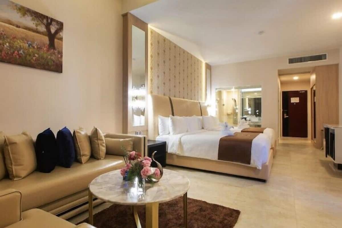 Ein modernes Hotelzimmer mit einer Couch, einem runden Couchtisch mit Blumen, einem großen Bett mit Kopfteil und einem Spiegel.