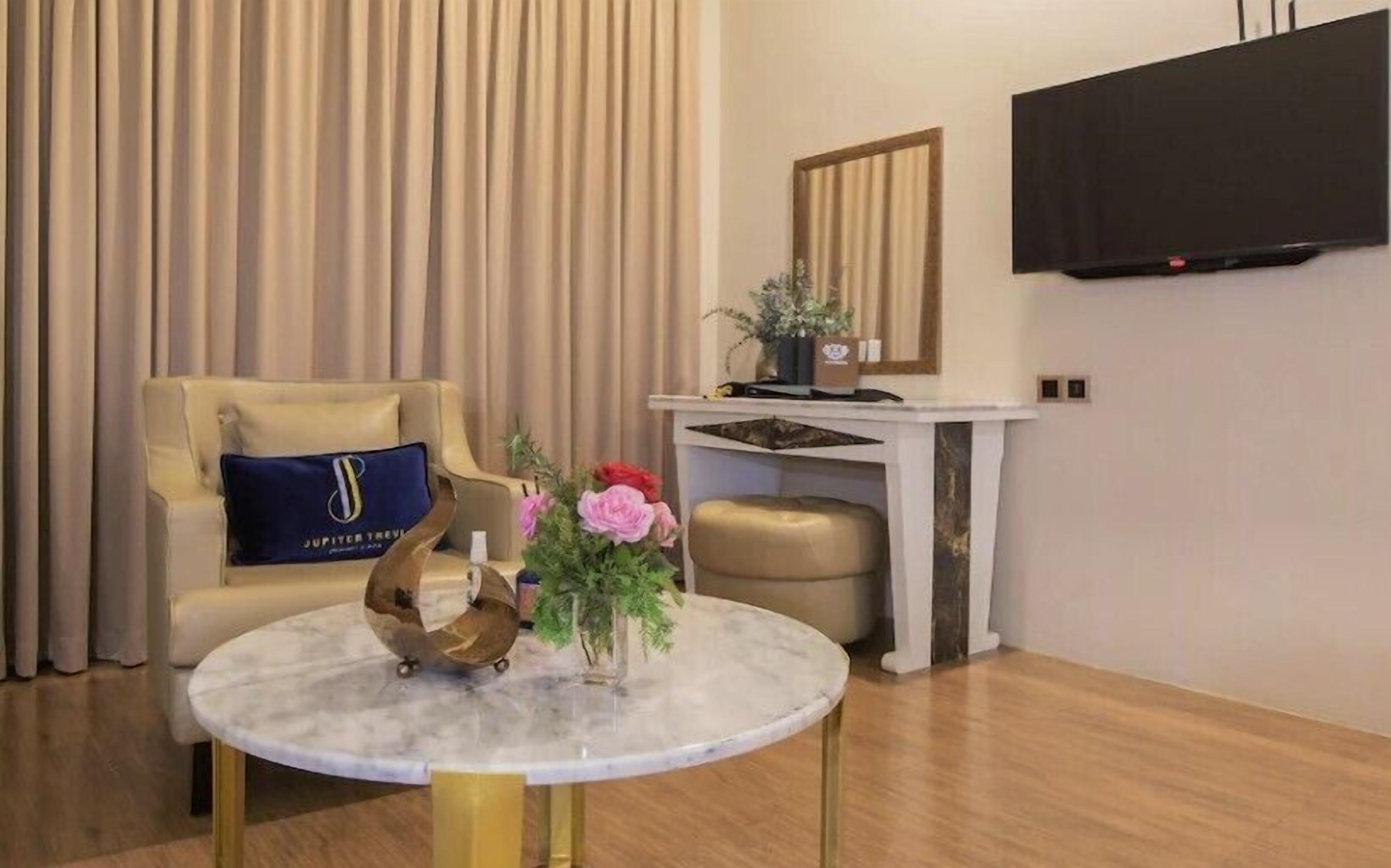 A modern living room with a round marble table, a beige sofa with a blue pillow, a small stool, a white console table with a mirror, and a flat-screen TV mounted on the wall.