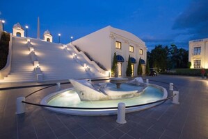 Un edificio blanco con una fuente en primer plano, rodeado de una zona pavimentada y escalones que lo llevan hacia arriba.