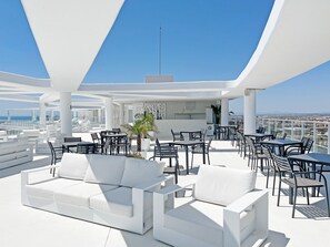 A rooftop terrace with white furniture, a bar area, and a view of the city. - Global Vacacional: Stunning apartment with private pool and terrace overlooking the sea (Canet d´en berenguer)