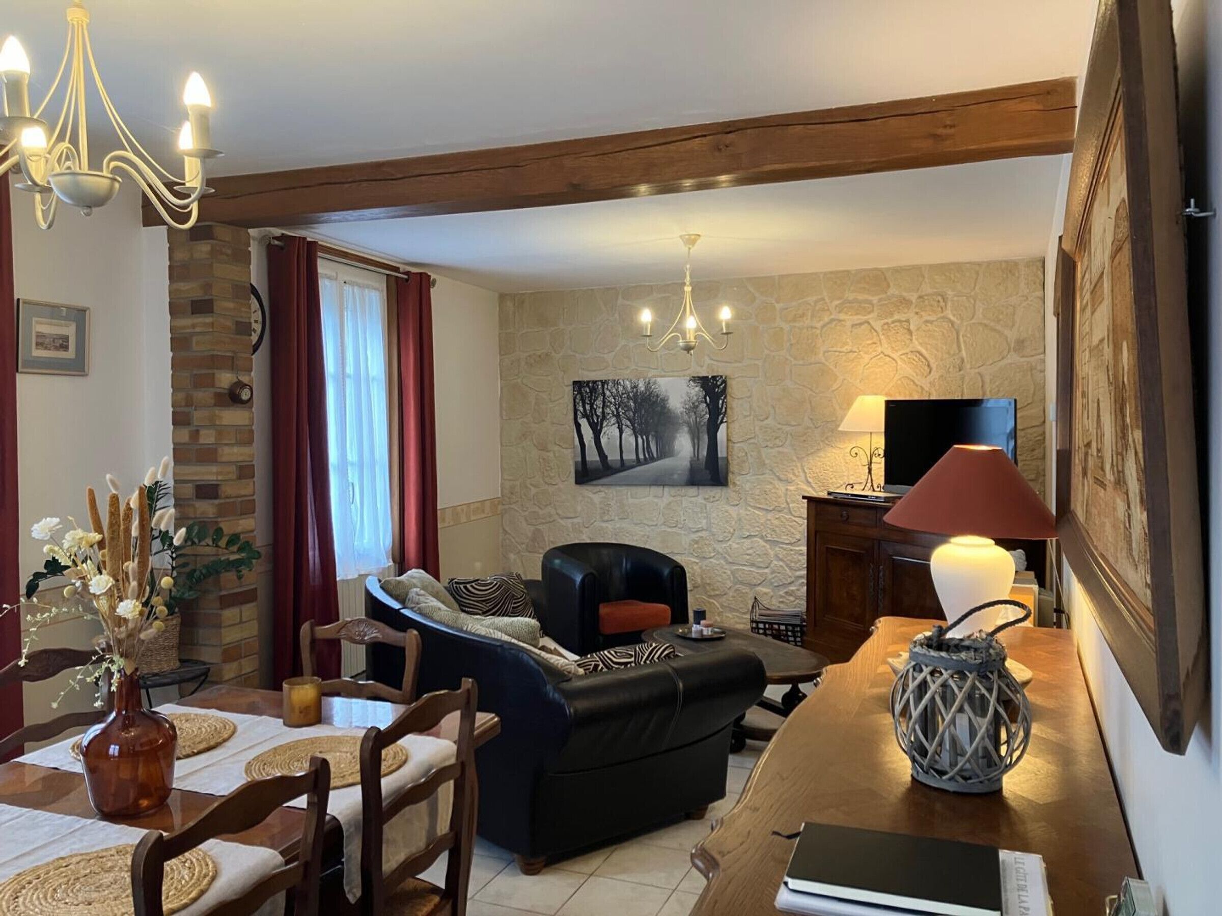 Un salon chaleureux avec un mur en pierre, une cheminée, un canapé en cuir noir, une table à manger avec des sets de table et un lustre.