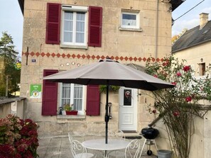 A stone building with red shutters, a white door, and a small window. There is a table with a metal chair and a large umbrella. - Charming 2-Bedroom Stone House with Garden, Near Pierrefonds Castle (Cuise-la-Motte)