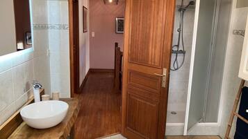 A bathroom with a wooden vanity, a large white basin, a shower with a glass door, and a wooden door leading to a hallway.
