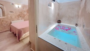 A room with a large bathtub filled with water and rose petals, a pink towel, and a small round object on the edge.