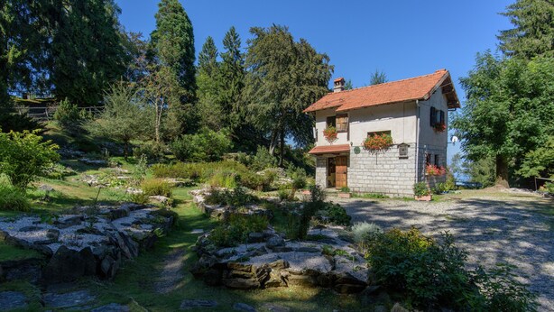 Uma casa de pedra com telhado de telhas vermelhas, rodeada por um jardim com pedras e vegetação.