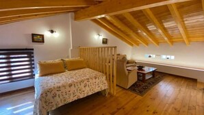 Ein gemütliches Schlafzimmer im Dachgeschoss mit Holzbalkendecke, einem Bett mit floralem Bettzeug, einem Schreibtisch und einem Fenster mit Jalousien.