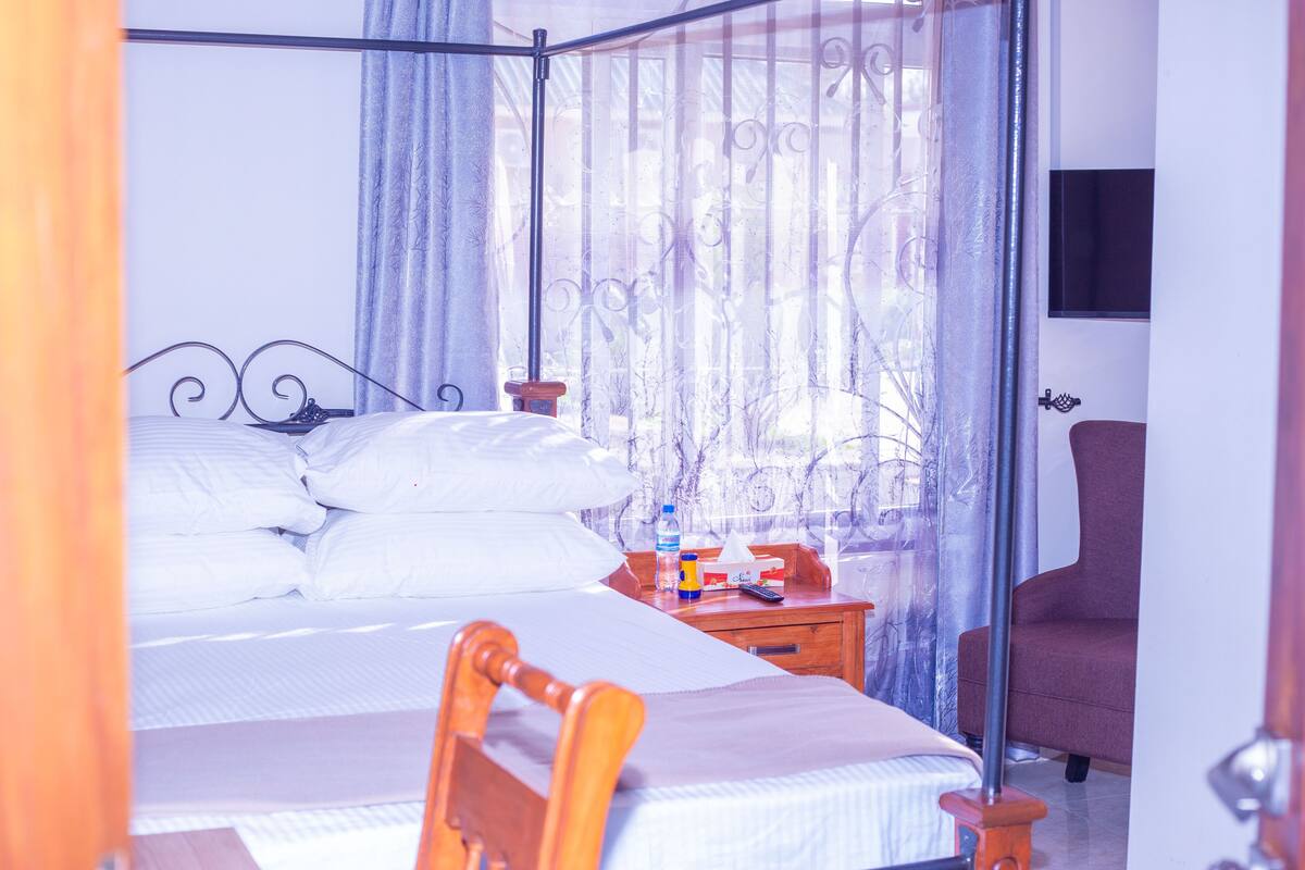 A four-poster bed with white linens, a wooden nightstand with a bottle and tissues, and a chair in a room with a view through a window with curtains.