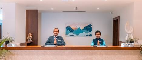 A reception desk with two individuals seated, a painting of mountains on the wall, and a reception sign.