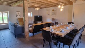 Ein Essbereich mit einem langen Holztisch, gedeckt für sechs Personen, umgeben von schwarzen Stühlen. Im Hintergrund befinden sich eine Couch und ein Fernseher.