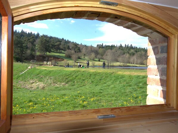 A view through an arched window of a grassy field with a lake, trees, and a wooden structure.