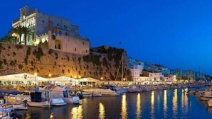 A coastal marina with boats docked, a castle-like building on a hill, and a row of buildings along the waterfront.
